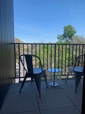 Ein Balkon mit zwei Stühlen und einem kleinen Tisch mit Blick auf eine Straße und Bäume.