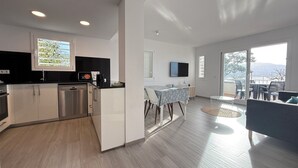 Um apartamento moderno com cozinha, sala de jantar e sala de estar. Possui uma grande janela com vista, televisão de tela plana e varanda com móveis para área externa.