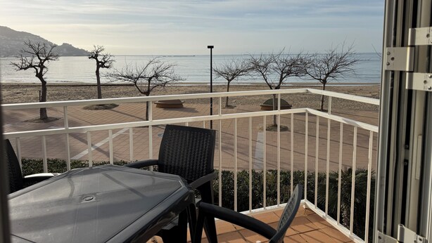 Varanda com mesa e cadeiras, vista para uma praia com árvores e céu claro.
