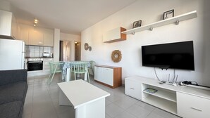 Un salon moderne doté d’un téléviseur à écran plat, d’un coin repas avec une table et des chaises, et d’une cuisine en arrière-plan.
