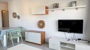 Un salon moderne avec un téléviseur, des meubles blancs, une table à manger avec des chaises et des œuvres d’art décoratives au mur.