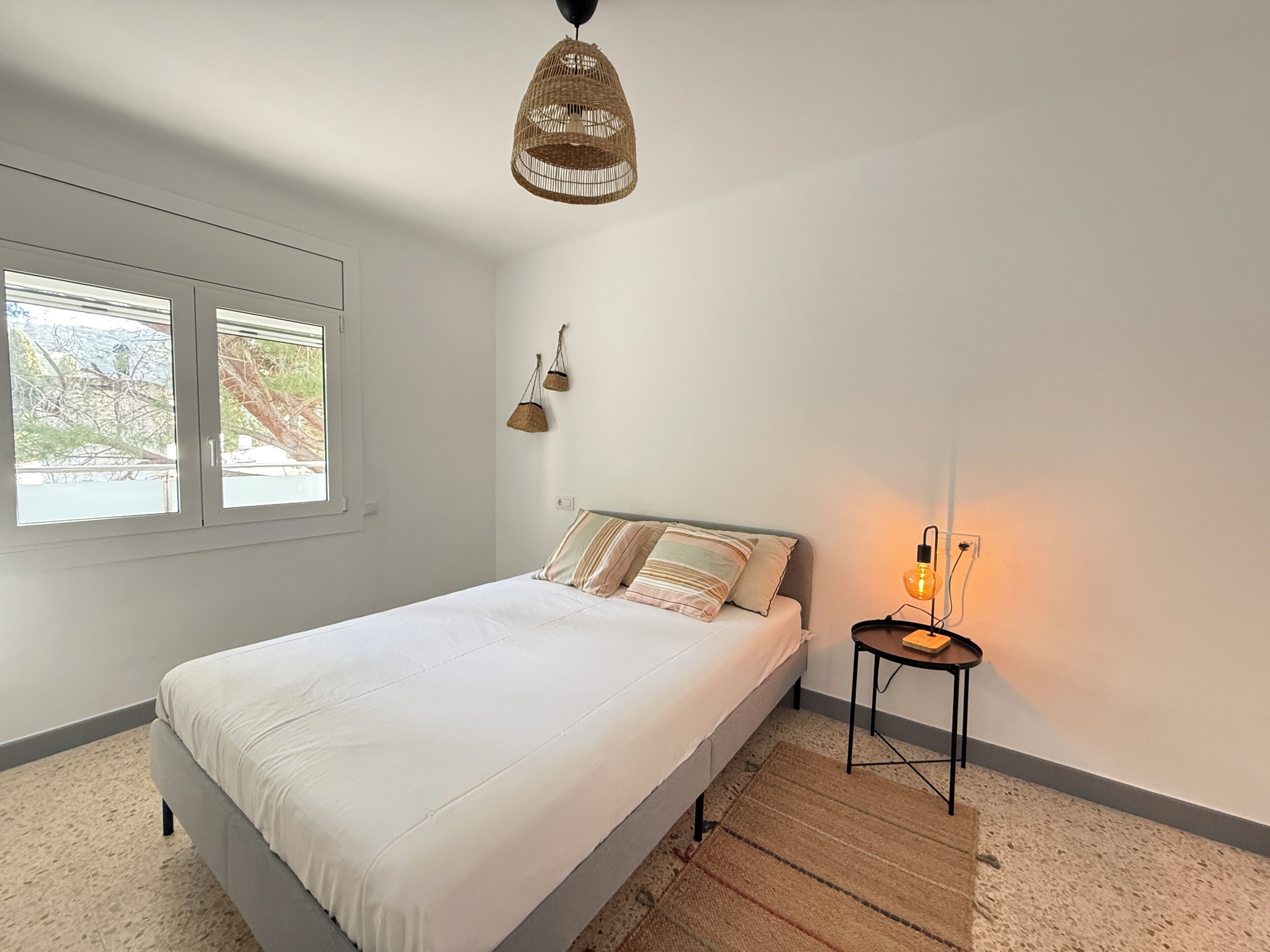 A bedroom with a bed, a bedside table with a lamp, a window, and a hanging light fixture.