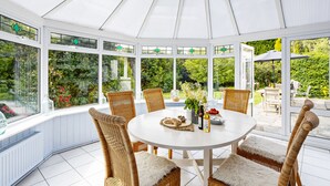 A conservatory with a round table, wicker chairs, and a view of greenery. - Blue Haze (St Austell)