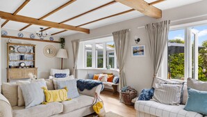 A cozy living room with a high ceiling, wooden beams, a sofa set, a side table, a lamp, and a window with curtains. - Blue Haze (St Austell)