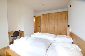 A bedroom with a wooden wardrobe, a desk with a chair, and a bed with white bedding.