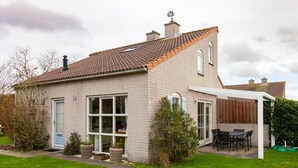 Une maison à deux étages, avec un toit en tuiles, un patio couvert équipé d’une table et de chaises, et une pelouse bien entretenue.