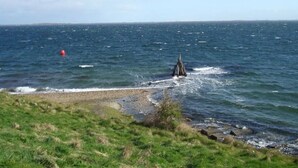 Un paysage côtier avec une zone herbeuse qui descend vers la mer, une bouée et une structure dans l’eau.