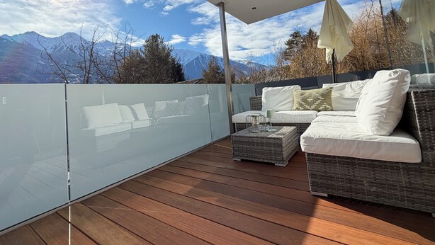 Een balkon met een houten vloer, een glazen hekwerk en een bank met kussens.