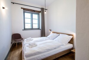 Un lit simple avec du linge de lit blanc, une tête de lit en bois, une fenêtre avec des rideaux, une chaise marron et un luminaire fixé au mur.