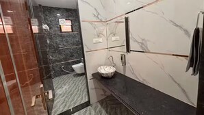 Un baño moderno con cabina de ducha de vidrio, pared con azulejos de mármol, encimera negra y lavamanos blanco.