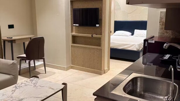 Habitación de hotel con cama, televisión montada en un panel de madera, escritorio con silla, mesita de centro de mármol y una zona de cocina con lavabo.