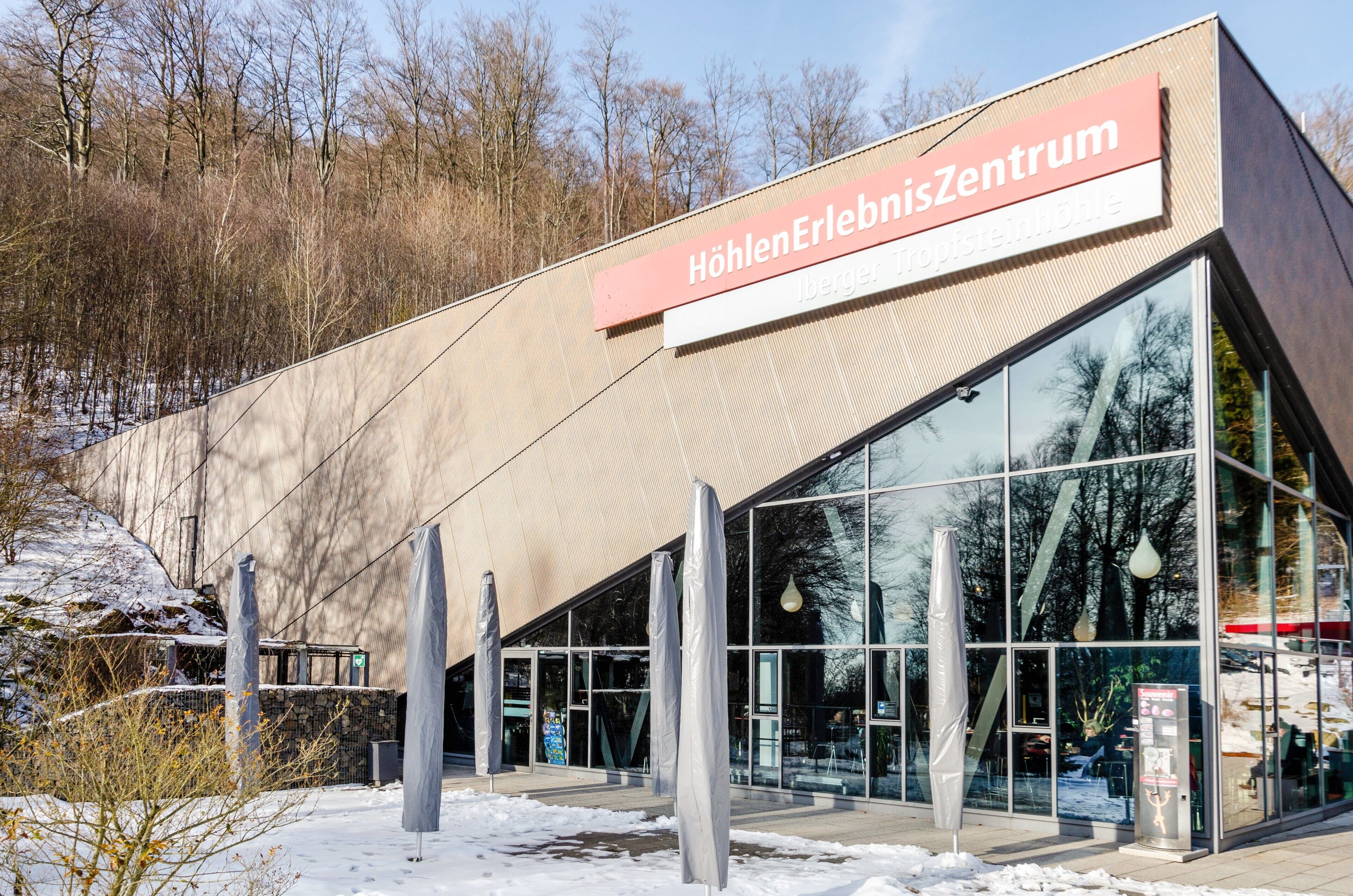 A modern building with a large glass facade, a prominent sign reading 'HöhlenErlebnisZentrum,' and a snow-covered ground.