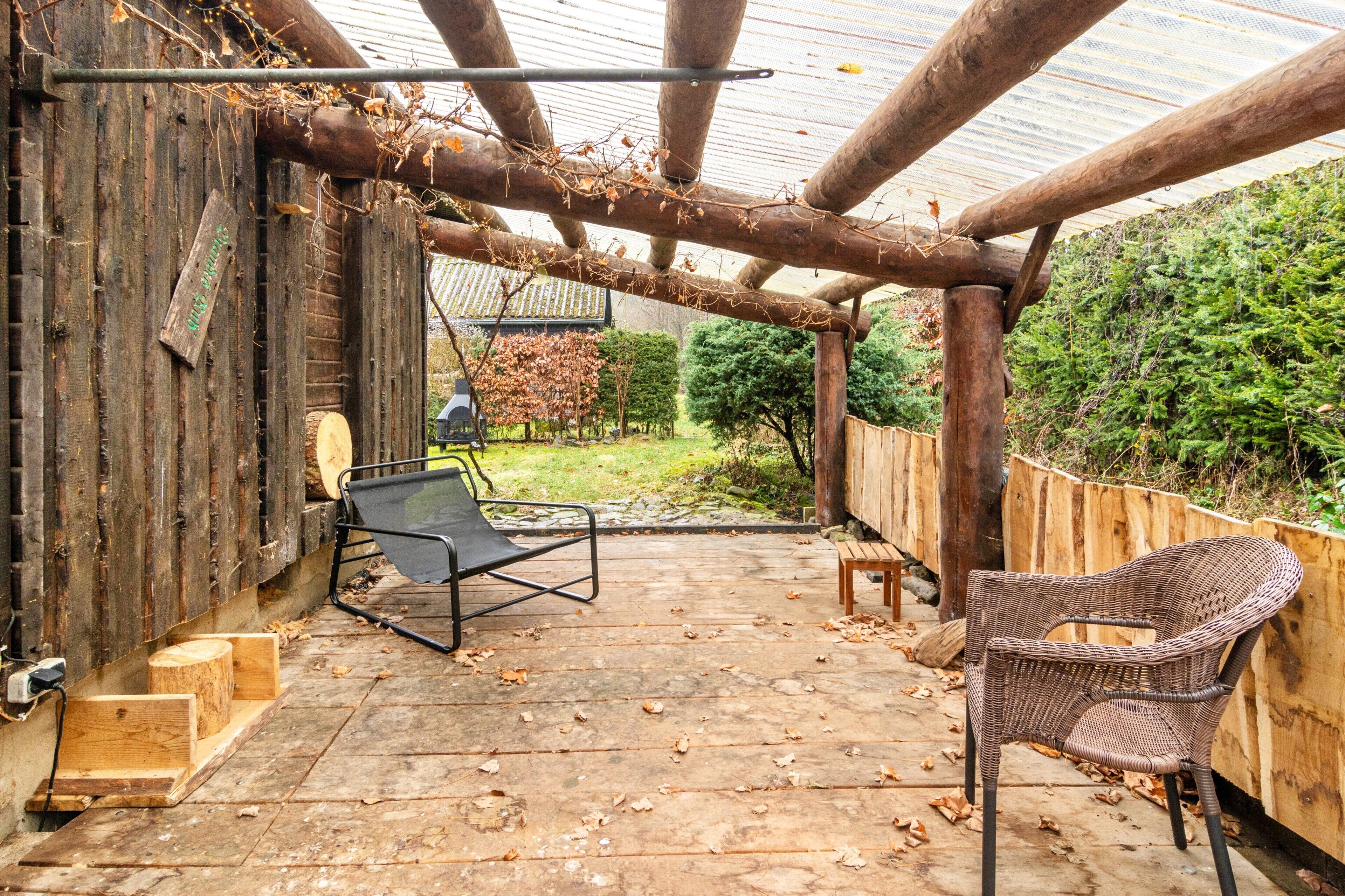 Een houten terras met een pergola, twee rieten stoelen en een kleine tafel.