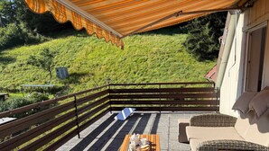 Un balcón con mesa de madera, muebles de mimbre y vistas a una ladera verde.