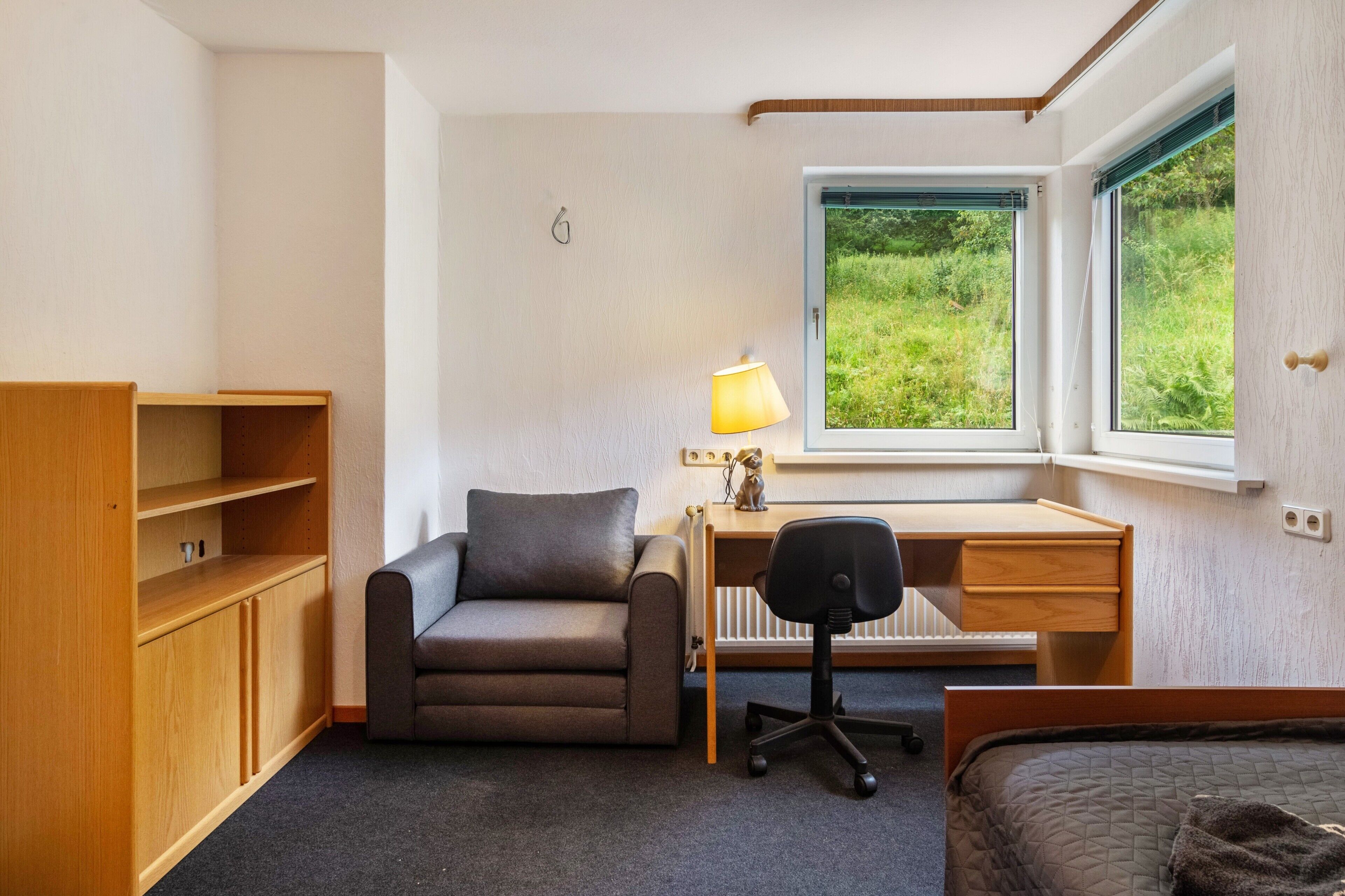Ein gemütlicher Raum mit Bett, Schreibtisch, Sessel und einer Couch, mit Blick auf Grünflächen.