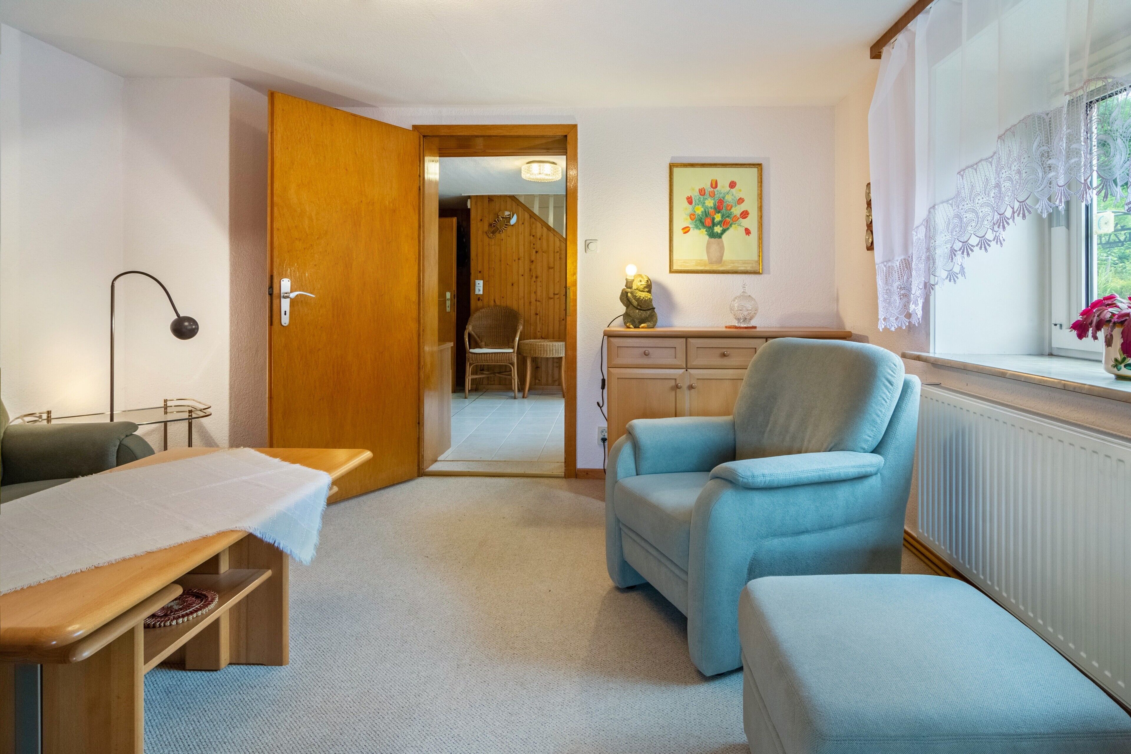 Une pièce avec une porte en bois, une petite table recouverte d’une nappe blanche, un fauteuil bleu, une chaise en osier, une commode ornée d’un vase et un tableau représentant des fleurs.