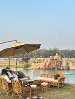 A serene outdoor seating area with wicker furniture and a large umbrella, overlooking a pond with a Buddha statue and a rock formation.