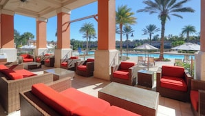 Zona relax esterna coperta con mobili in vimini e cuscini rossi, con vista su piscina e palme.