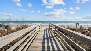 Une promenade en bois menant à une plage, avec un ciel dégagé et une vue sur l’océan.
