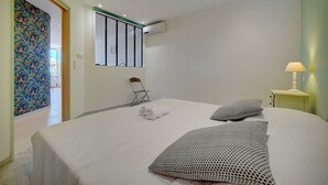 Una cama bien hecha con sábanas blancas y almohadas estampadas, una mesita de noche con lámpara y un equipo de aire acondicionado en la pared.