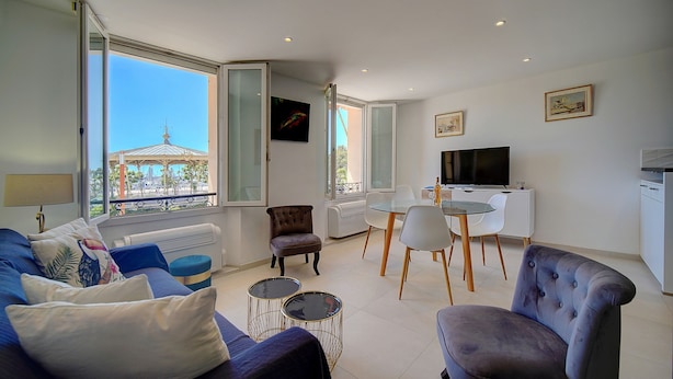 Una sala de estar moderna con un sofá, sillones, una mesa de comedor y una televisión. Amplias ventanas ofrecen vistas a un edificio con una estructura en forma de cúpula.