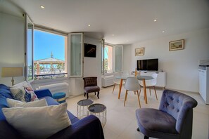 Un salon moderne comprenant un canapé, des fauteuils, une table à manger et un téléviseur. De grandes fenêtres offrent une vue sur un bâtiment doté d’une structure en forme de dôme.