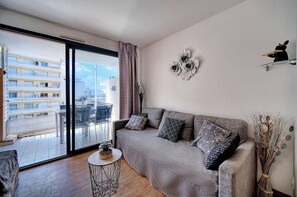 Un salon moderne avec un canapé gris, des coussins à motifs, une petite table ronde agrémentée d’une plante, et une vue sur un balcon aménagé avec du mobilier d’extérieur.