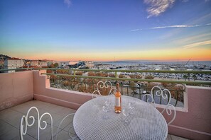 Un balcon avec une table ronde et des chaises, une bouteille de vin, et une vue sur un port de plaisance et le paysage urbain.
