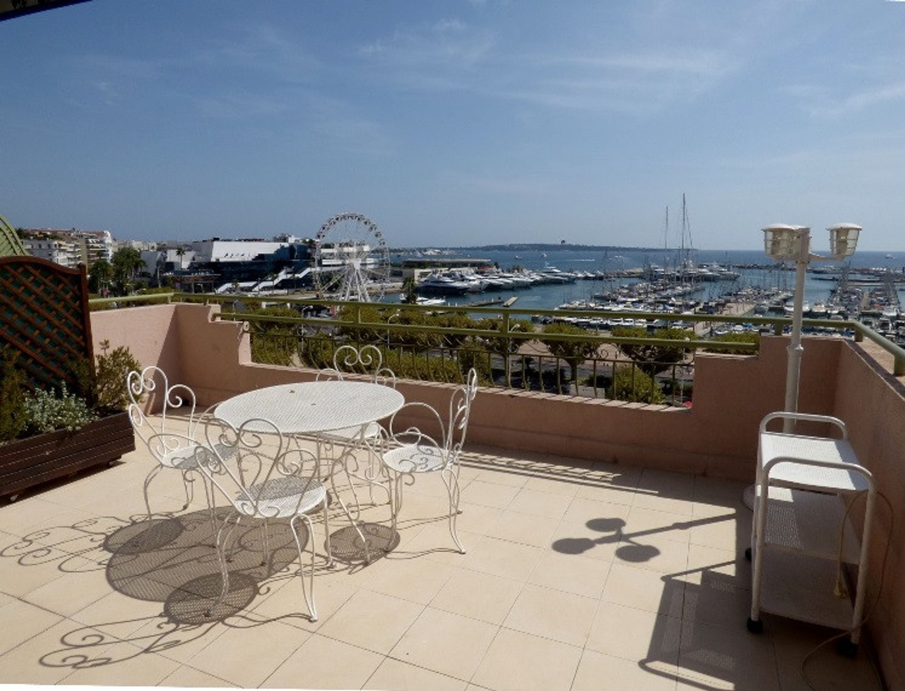 A terrace with a table and chairs, overlooking a marina with boats and a Ferris wheel.