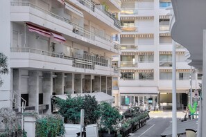 Ein mehrstöckiges Wohngebäude mit Balkonen, einem überdachten Eingang und Blick auf die Straße mit parkenden Fahrzeugen und Grünflächen.