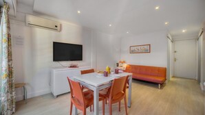 Un salon moderne avec une table à manger, des chaises orange, un canapé blanc, un téléviseur à écran plat et un tableau accroché au mur.