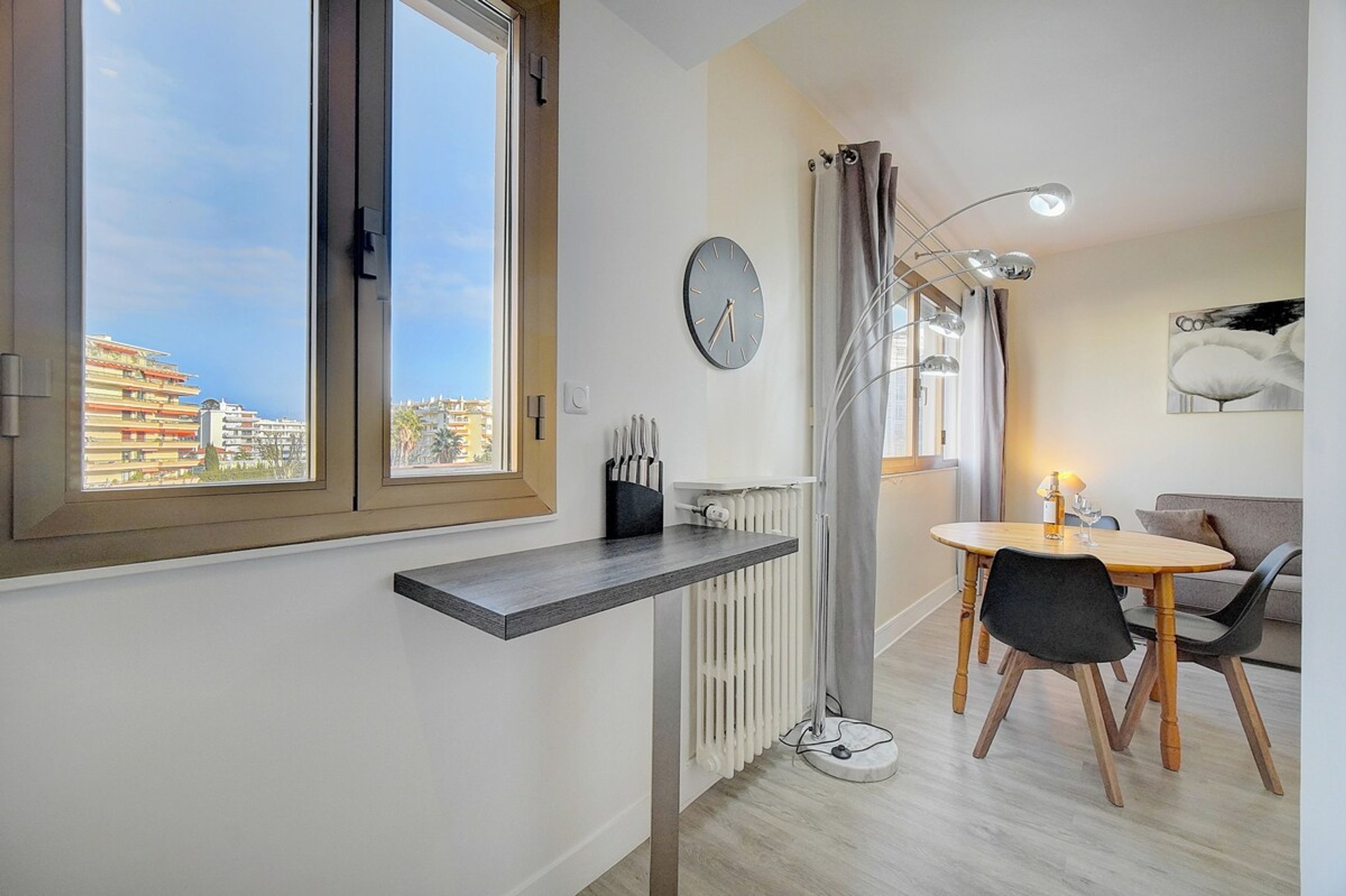 A modern living room with a dining area, large windows, a radiator, and a clock on the wall.