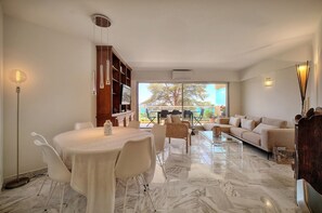 A spacious living room with a marble floor, a dining area with a round table, and a large window offering a view of the outdoors. - 3 pi ces Terrasse vue mer et Parking A2b232 (Cannes)