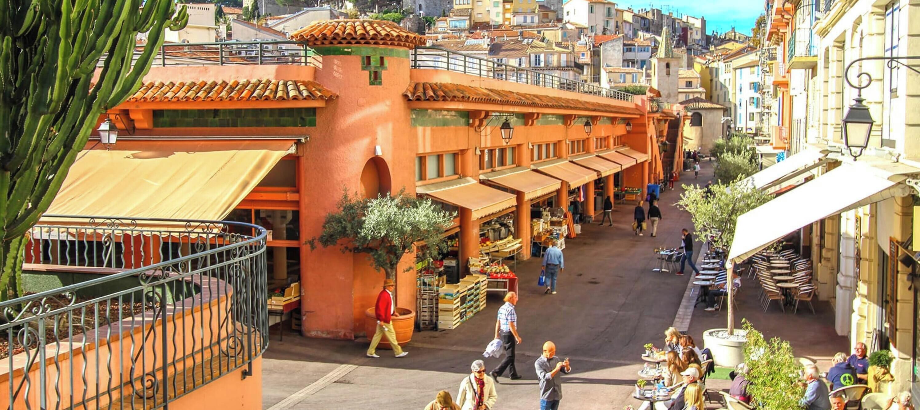 Une rue animée, avec des terrasses et un espace réservé à un marché, bordée de bâtiments à l’architecture traditionnelle.