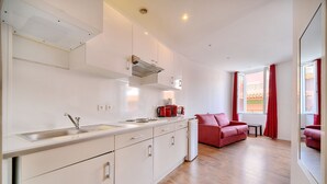 A modern kitchen with white cabinets, a stainless steel sink, and a red coffee machine. There is a red sofa in a living area with a large window and red curtains. - Charmant 2 Pieces Centre DE Cannes A1b209 (Cannes)