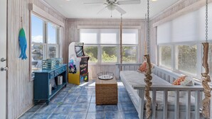 A sunroom with a ceiling fan, a large window, a play area with a game machine, and a wicker basket on the floor. - Sounds Right #214-Community pool, private boat dock, kayaks, fish, soundside (North Topsail Beach)