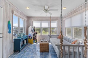 A sunroom with a ceiling fan, a large window, a play area with a game machine, and a wicker basket on the floor. - Sounds Right #214-Community pool, private boat dock, kayaks, fish, soundside (North Topsail Beach)