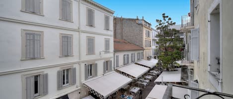 Rua estreita com mesas ao ar livre sob toldos, edifícios brancos com persianas e um prédio de pedra ao fundo.
