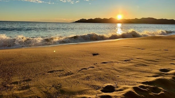Ein Strand mit Fußabdrücken, die zum Wasser führen, ein Sonnenuntergang im Hintergrund und eine entfernte Landmasse.