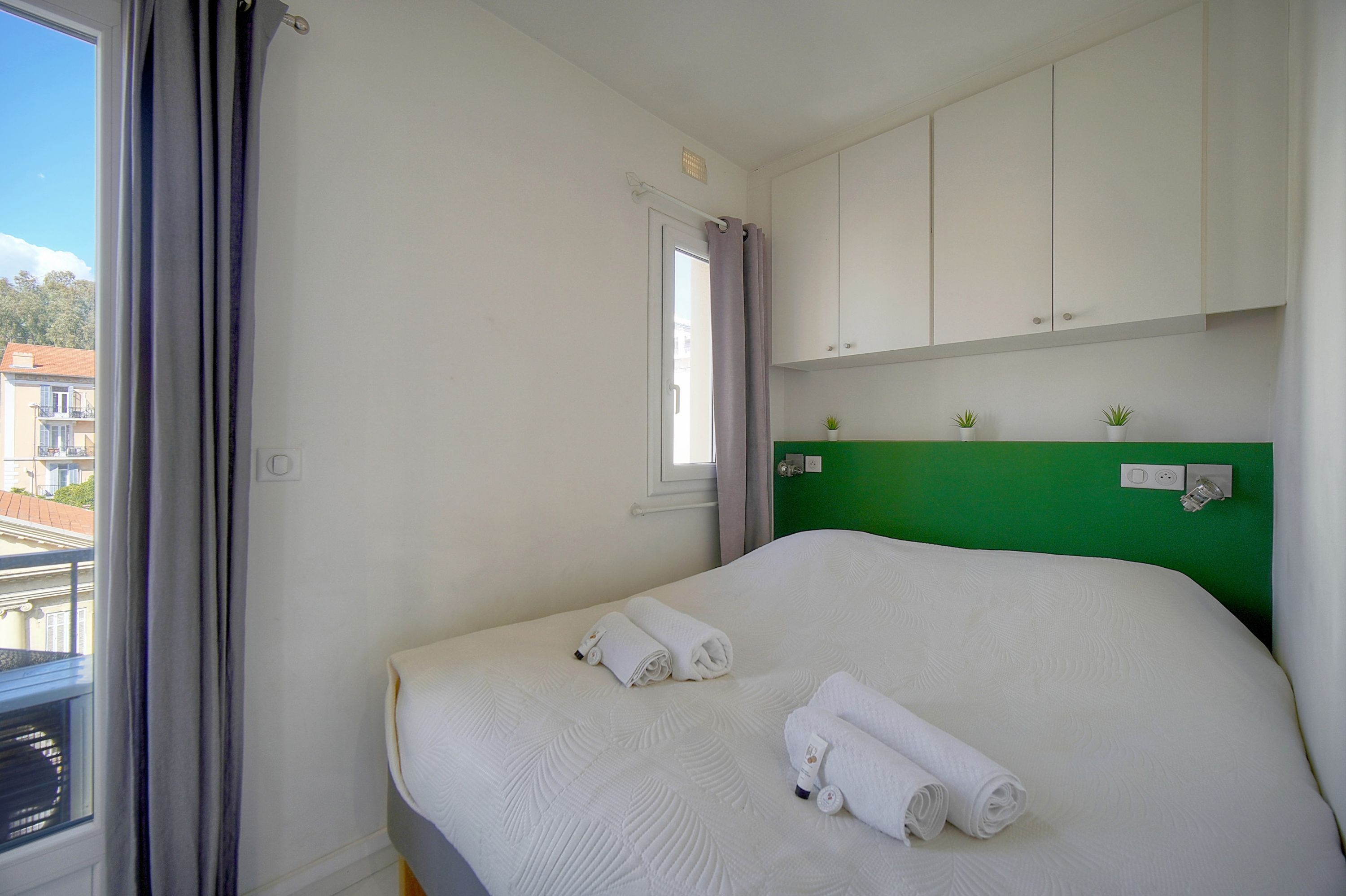 Una camera d'albergo con un letto, una finestra con tende, una testiera verde e una piccola pianta sulla testiera.