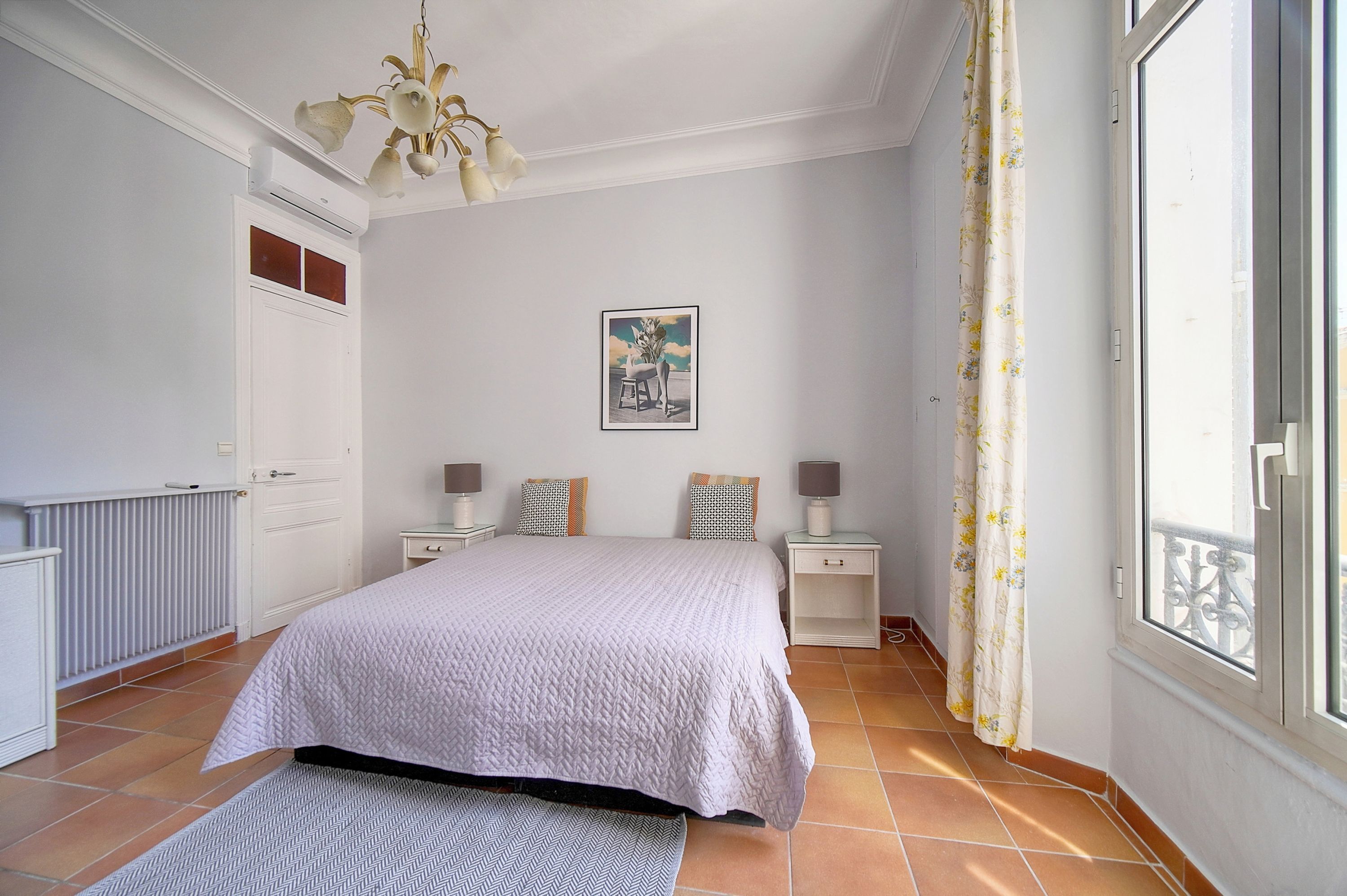 Une chambre à coucher avec un grand lit, deux tables de chevet avec des lampes, un radiateur, une fenêtre avec des rideaux et un luminaire au plafond.