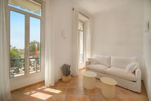Habitación luminosa con un sofá blanco, dos mesitas redondas y un balcón con vistas.