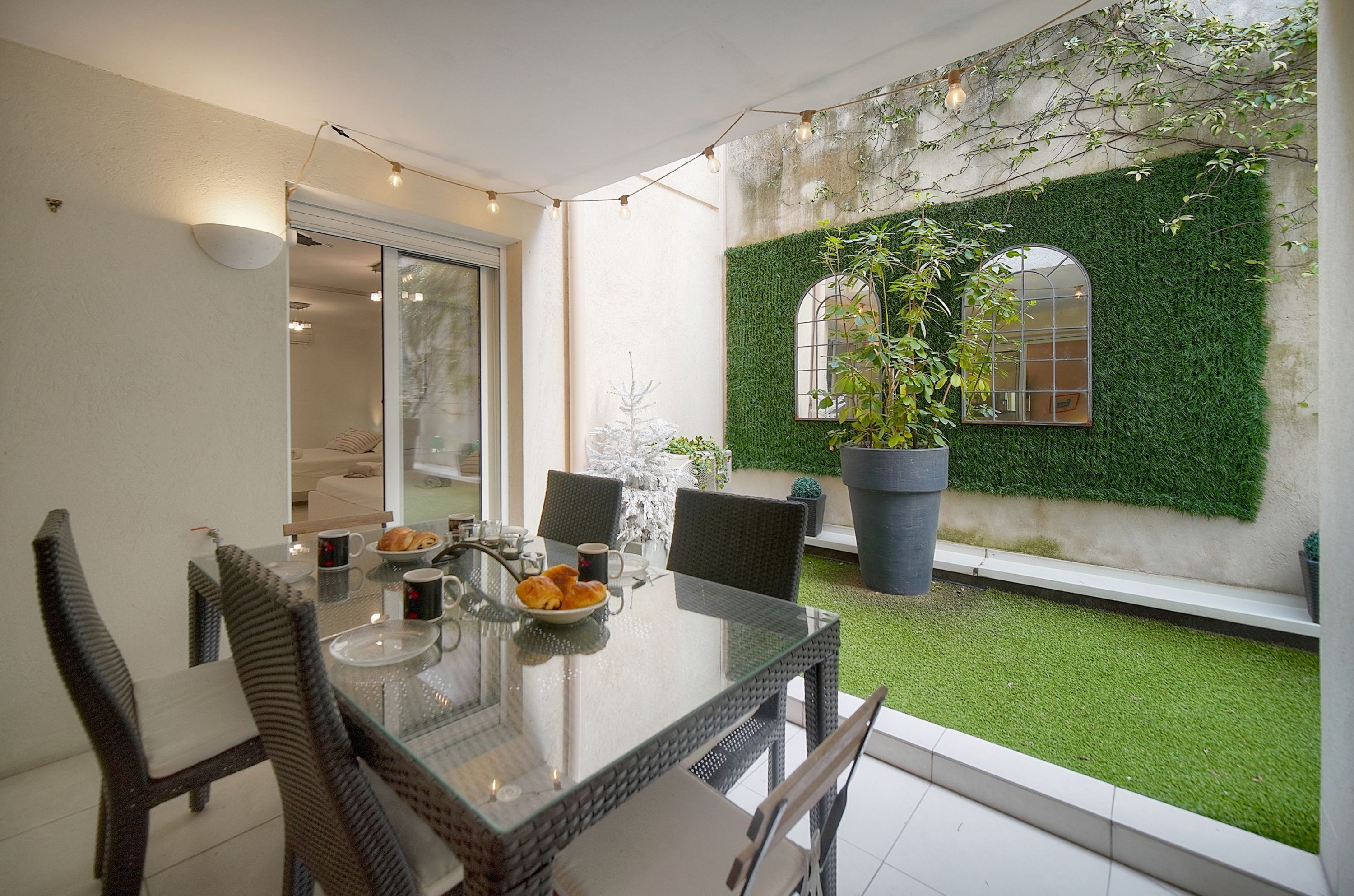 A dining area with a glass table, wicker chairs, and a view of a green wall and garden.