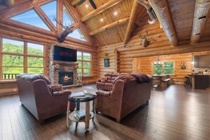 A log cabin interior with a fireplace, leather sofas, a television, and large windows overlooking a forest. - Beautiful Log Cabin-Easy Access-Heated Pool-HotTub-GameRoom-Theatre-Pet Friendly (Sevierville)