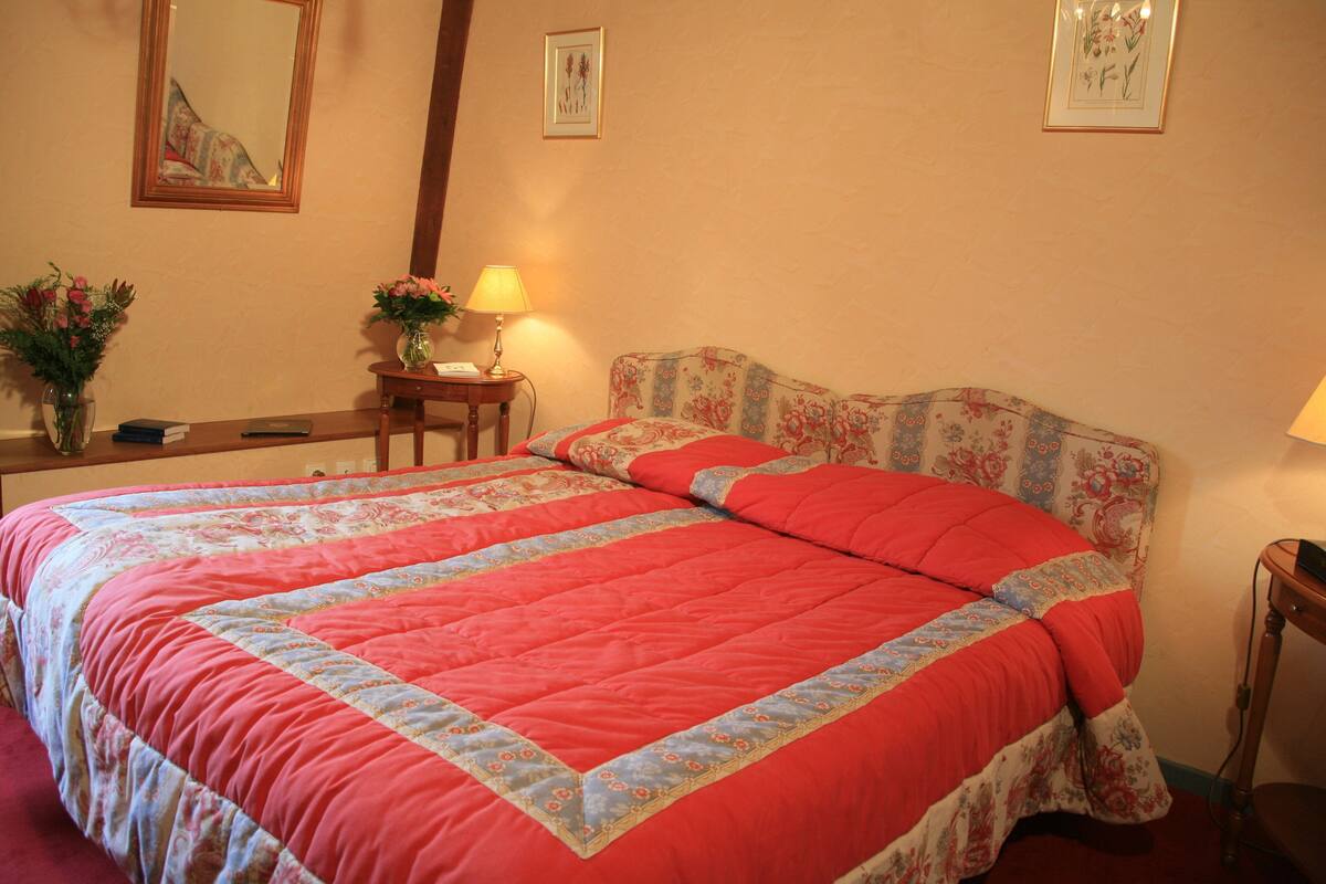 Ein ordentlich bezogenes Bett mit einem blumengemusterten Kopfteil, ein Nachttisch mit Lampe und eine Vase mit Blumen.