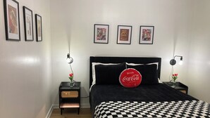 Ein Schlafzimmer mit einem schwarz-weiß karierten Überwurf, einem schwarzen Kopfteil und drei gerahmten Bildern an der Wand. Es gibt zwei Nachttische mit Lampen und ein rotes Coca-Cola Kissen auf dem Bett.