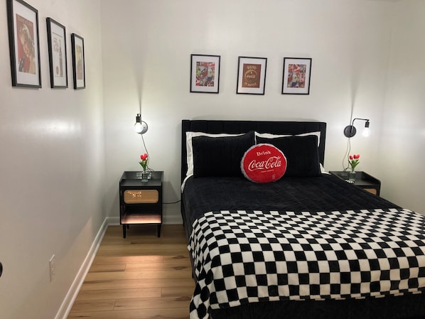 A bedroom with a black and white checkered bedspread, a black headboard, and three framed pictures on the wall. There are two bedside tables with lamps and a red Coca-Cola pillow on the bed.