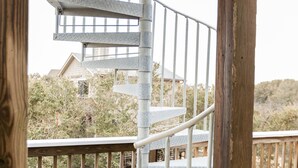 Eine Wendeltreppe mit Metallgeländern, einer Holzdeckkonstruktion und Blick auf ein Haus sowie Vegetation im Hintergrund.
