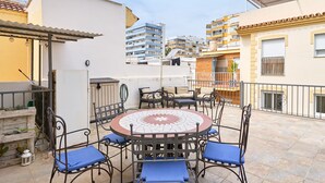 Eine Terrasse mit rundem Tisch und Stühlen, ein weißes Gebäude und ein mehrstöckiges Wohnhaus im Hintergrund.
