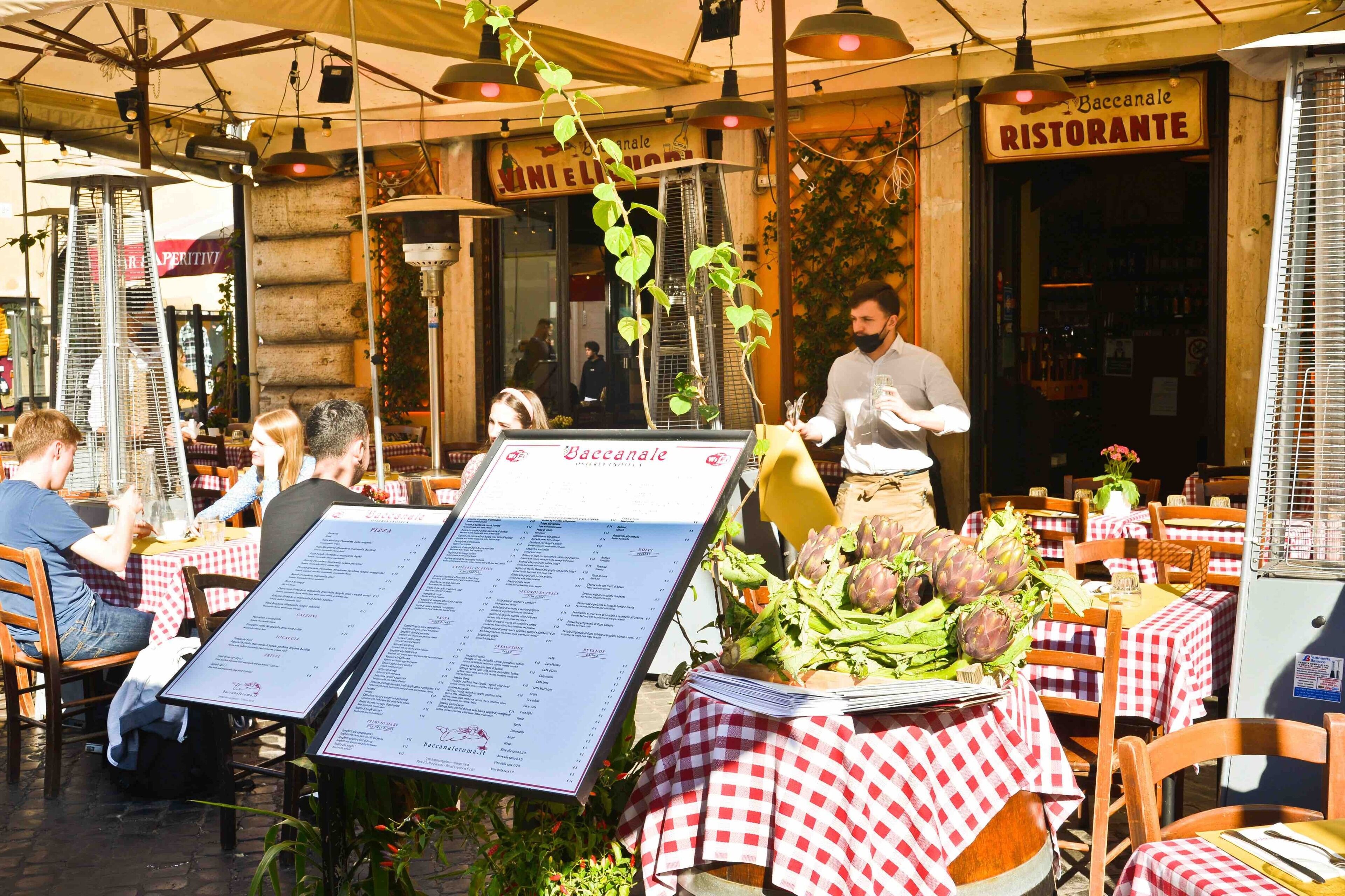 Un espace de restauration en plein air avec des tables recouvertes de nappes à carreaux rouges et blancs, un présentoir à menus et une variété de légumes frais exposés sur une table.